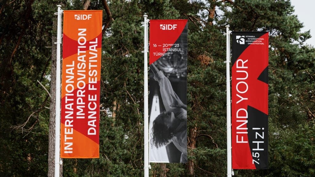 hree vertical outdoor banners promoting the Istanbul International Improvisation Dance Festival (IIDF), displayed against a backdrop of lush green trees. The left banner features bold orange and red text reading 'International Improvisation Dance Festival,' the center banner highlights the event dates, '16–20 May 2023, Istanbul, Turkey,' with an image of a dancer mid-performance, and the right banner displays the slogan 'Find Your 7.5 Hz!' in striking red and black.