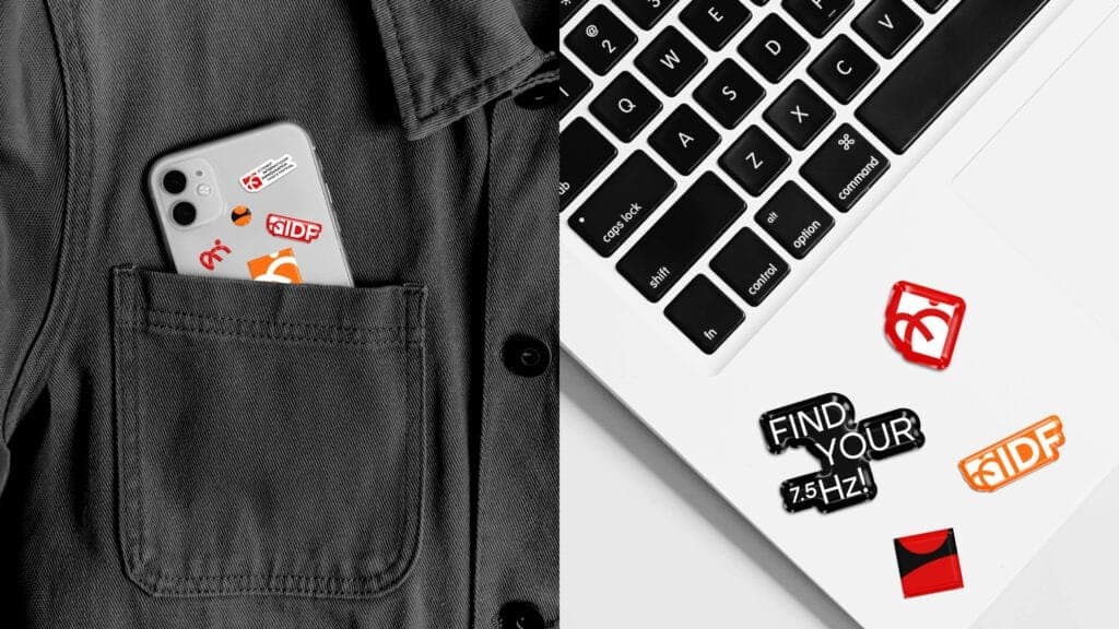 Stylish branding for the Istanbul International Improvisation Dance Festival (IIDF). On the left, a gray phone adorned with IIDF-themed stickers, tucked into a black jacket pocket. On the right, a laptop keyboard featuring bold and colorful IIDF stickers, including the slogans 'Find Your 7.5 Hz!' and the IIDF logo in red and orange, showcasing creative and modern promotional materials.